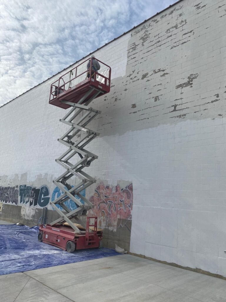 A person on a scissor lift against a large white brick wall, painting over graffiti in a commercial area. Below, a blue tarp is spread on the ground to catch any drips.