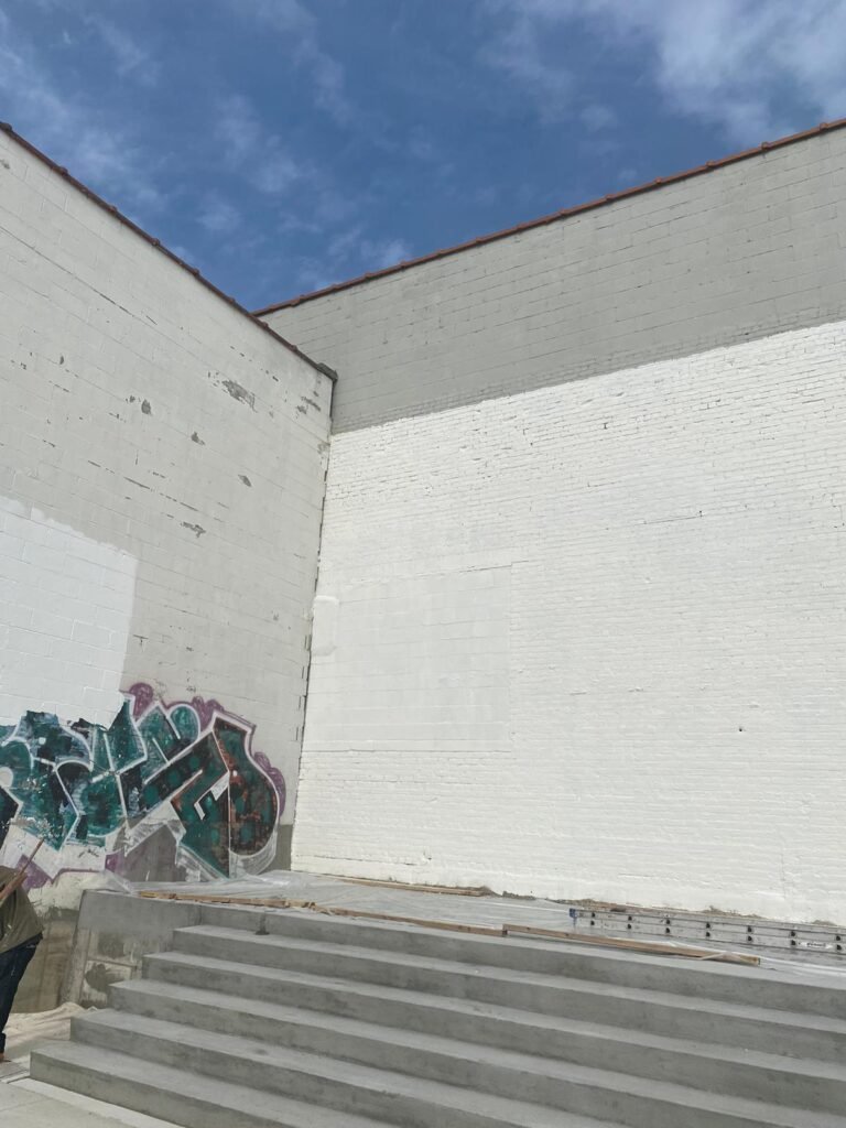 A broad, white-brick wall with a section partially painted over in the center. The adjacent wall on the left features graffiti in shades of green and purple. Concrete steps lead up to the walls, and the sky is clear with some scattered clouds.